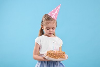 Photo of Birthday celebration. Cute little girl in party hat holding tasty cake with burning candles on light blue background