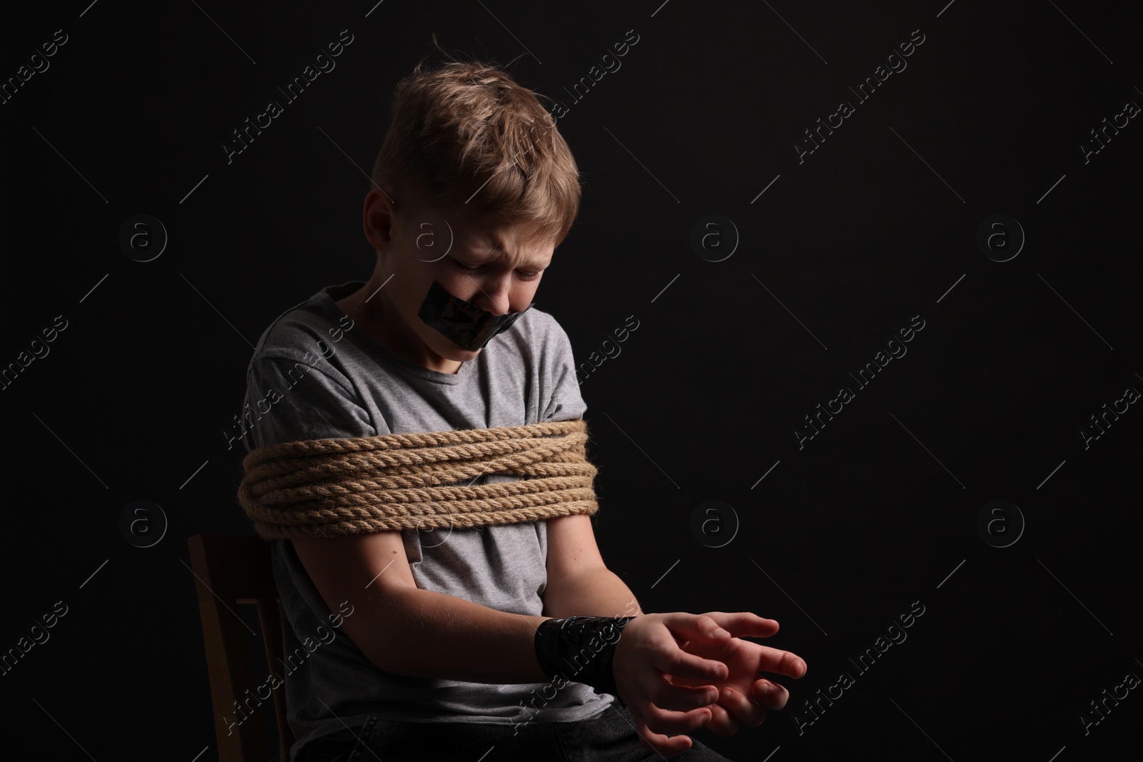 Photo of Little boy with taped mouth tied up and taken hostage against dark background. Space for text