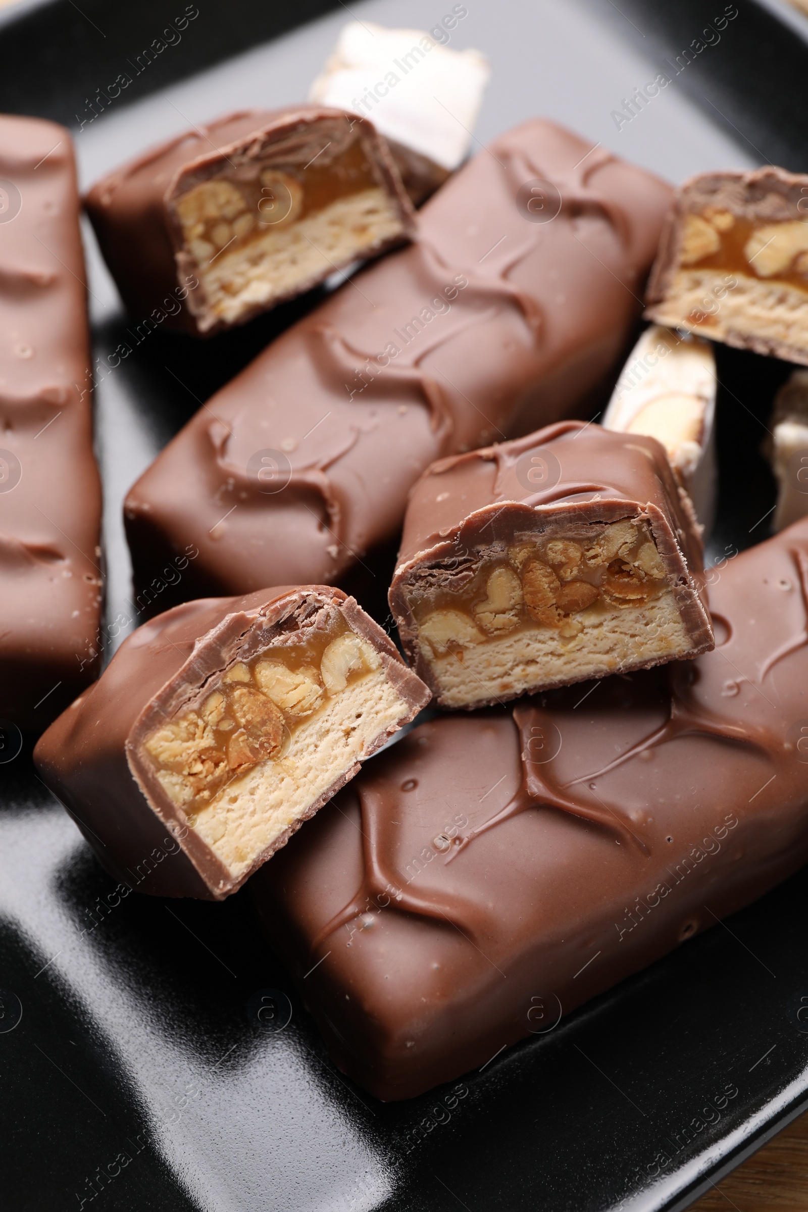 Photo of Tasty chocolate bars with nougat and nuts on black plate, closeup