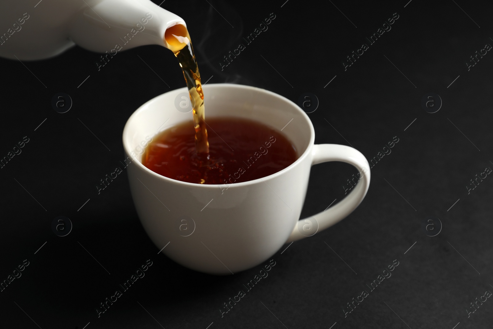 Photo of Pouring hot tea into white porcelain cup on dark background