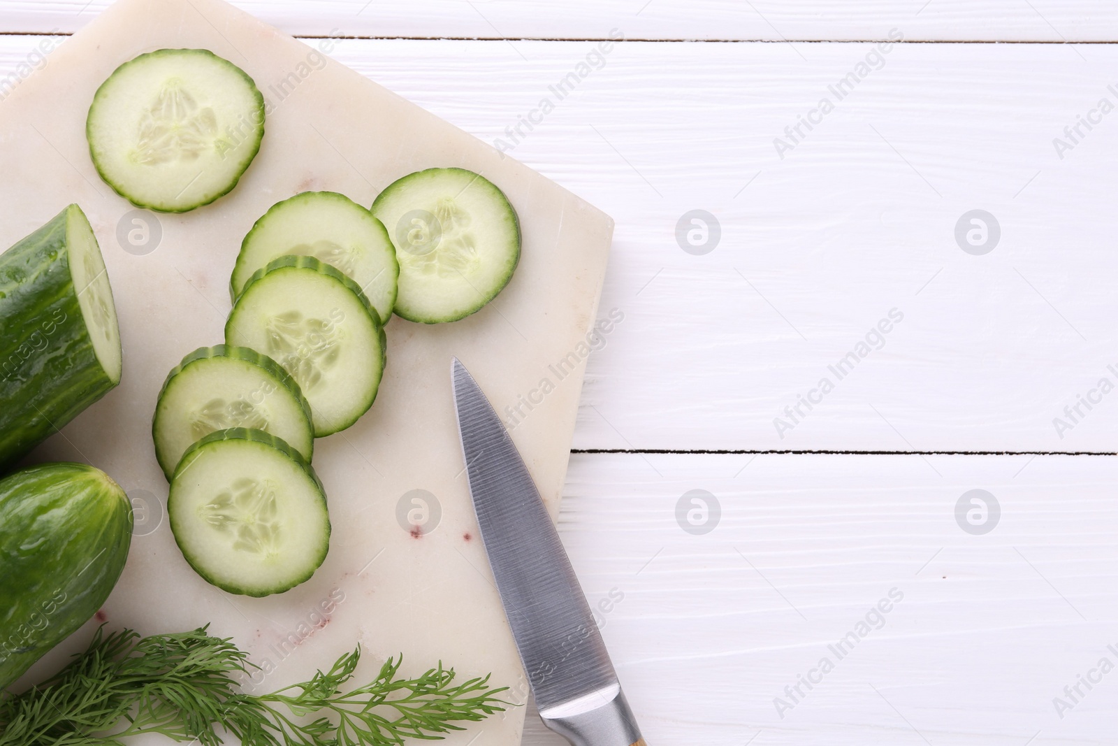 Photo of Cucumbers, dill, knife and marble cutting board on white wooden table, top view. Space for text