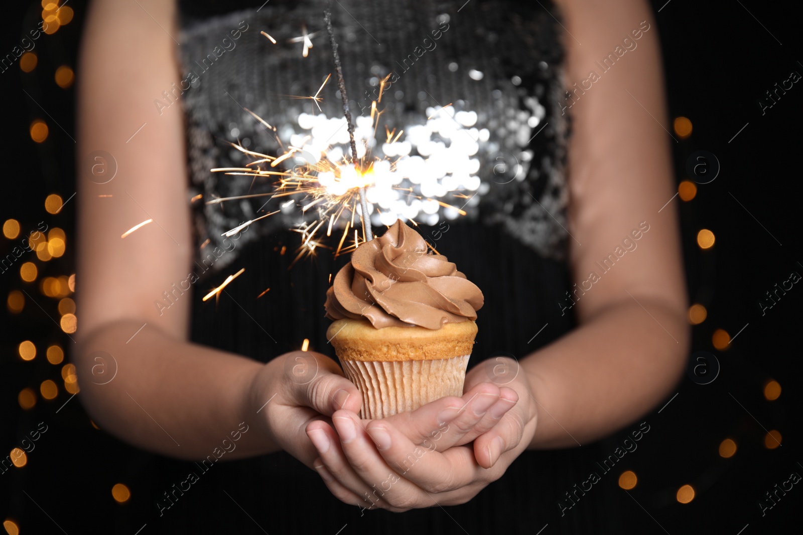 Photo of Woman holding birthday cupcake with sparkler on blurred background, closeup