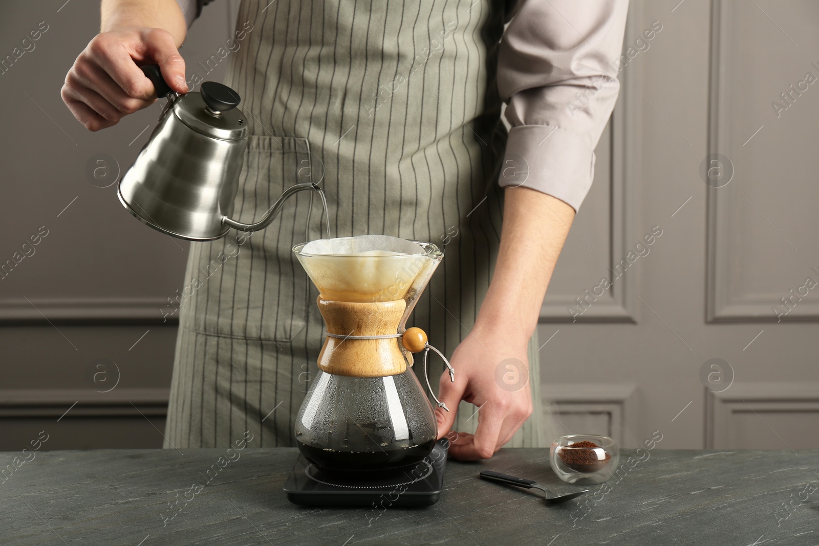 Photo of Man pouring hot water into glass chemex coffeemaker with paper filter and coffee at gray table, closeup
