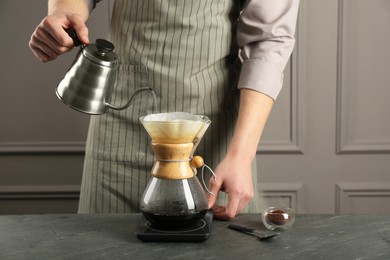 Man pouring hot water into glass chemex coffeemaker with paper filter and coffee at gray table, closeup