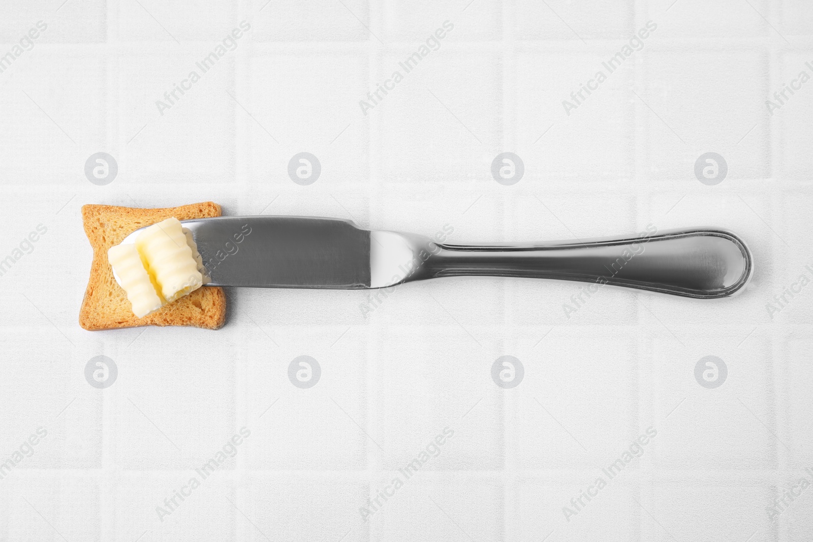 Photo of Tasty butter curl, knife and piece of dry bread on white tiled table, top view