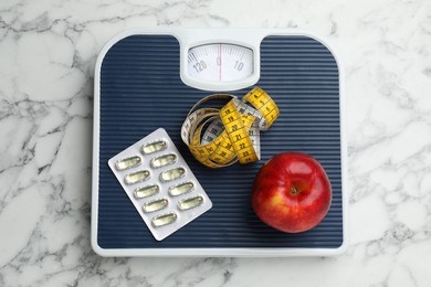 Photo of Scales with oil capsules, apple and measuring tape on white marble table, top view. Weight loss