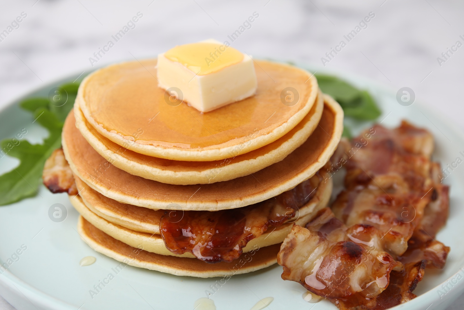 Photo of Tasty pancakes with butter, fried bacon and fresh arugula on white marble table, closeup