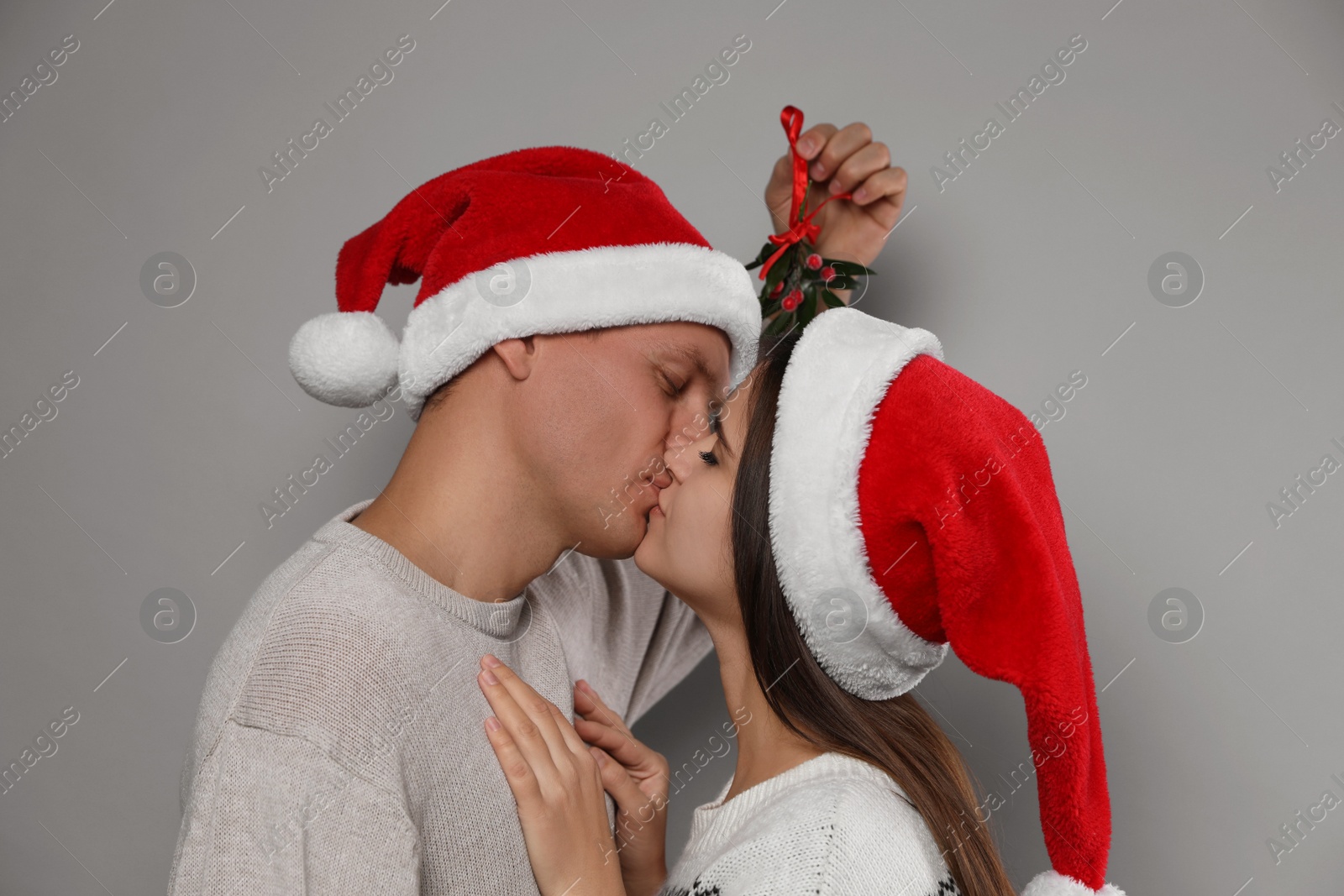 Photo of Happy couple in Santa hats kissing under mistletoe bunch on grey background