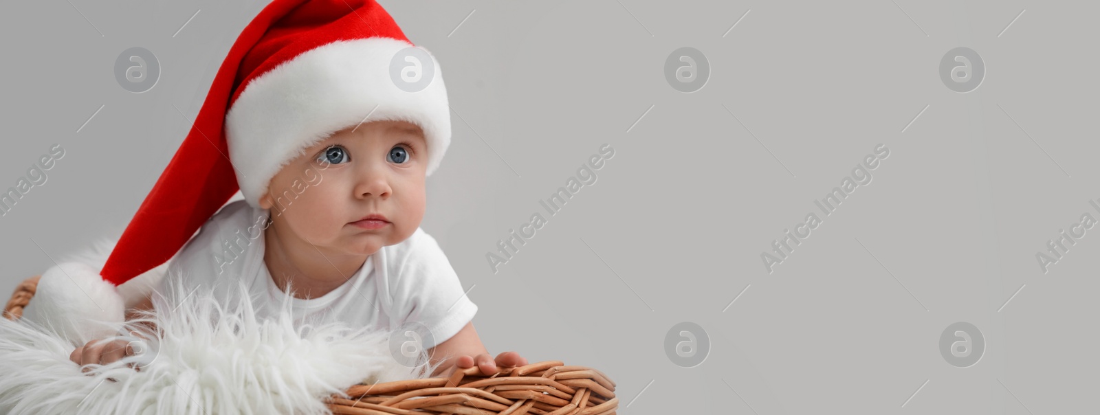Image of Cute baby wearing Santa hat in wicker basket on light grey background, banner design with space for text. Christmas celebration