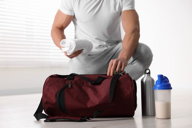 Young man putting towel into bag indoors, focus on protein shake and bottle of water