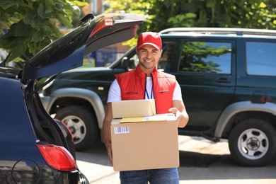 Young courier holding parcels near delivery car outdoors