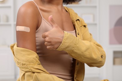 Photo of Young woman with adhesive bandage on her arm after vaccination showing thumb up indoors, closeup