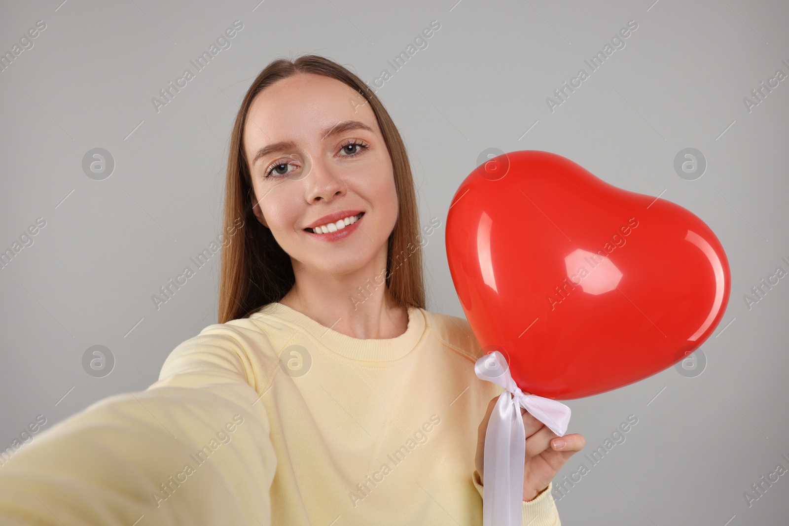 Photo of Happy young woman holding red heart shaped balloon and taking selfie on light grey background