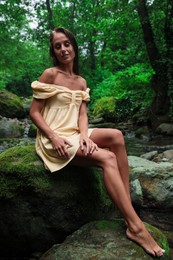 Photo of Beautiful young woman in dress sitting on rock near mountain river outdoors