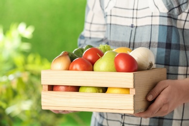Photo of Woman holding wooden crate filled with fresh vegetables and fruits outdoors, closeup