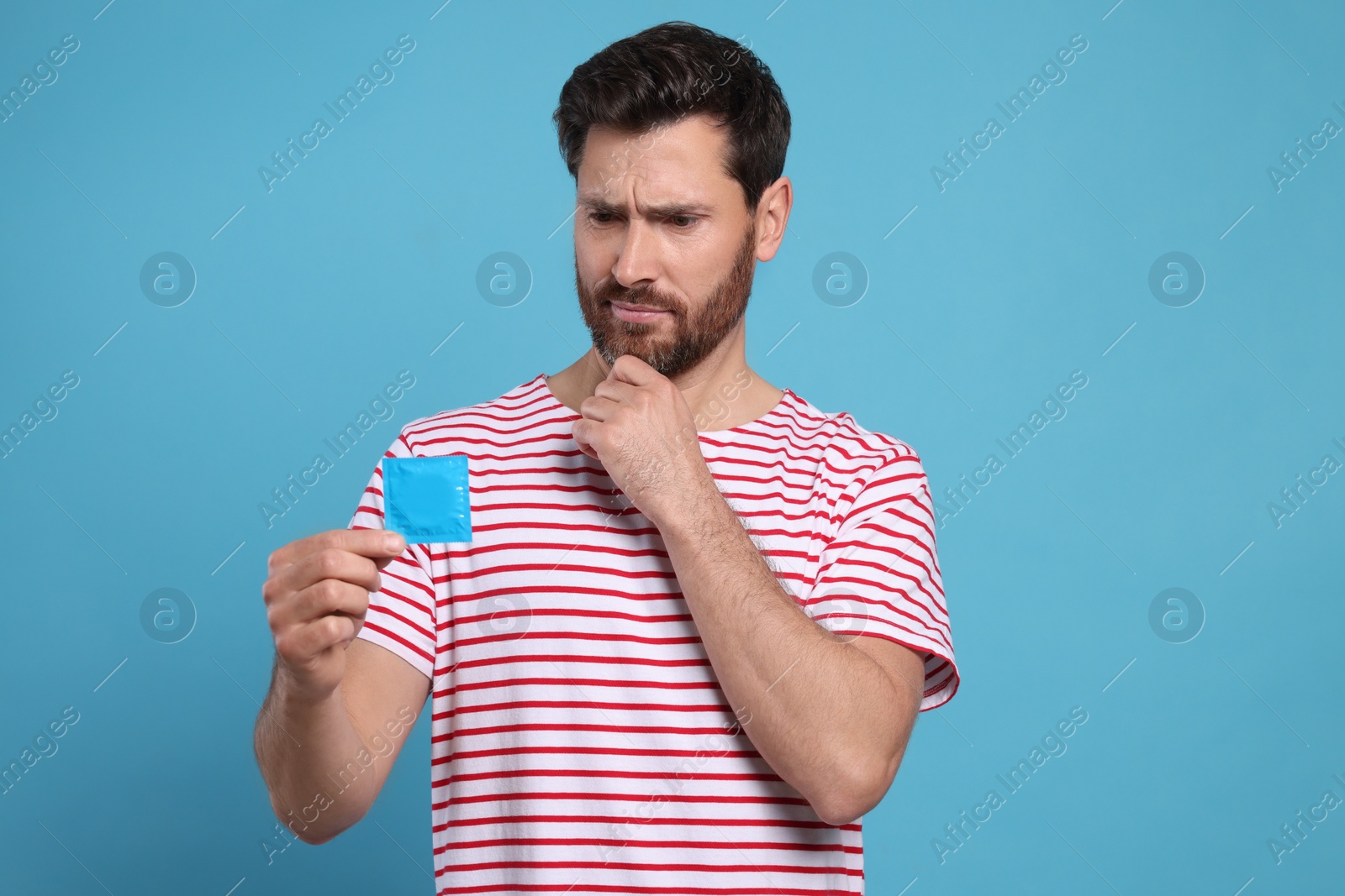 Photo of Confused man holding condom on light blue background. Safe sex