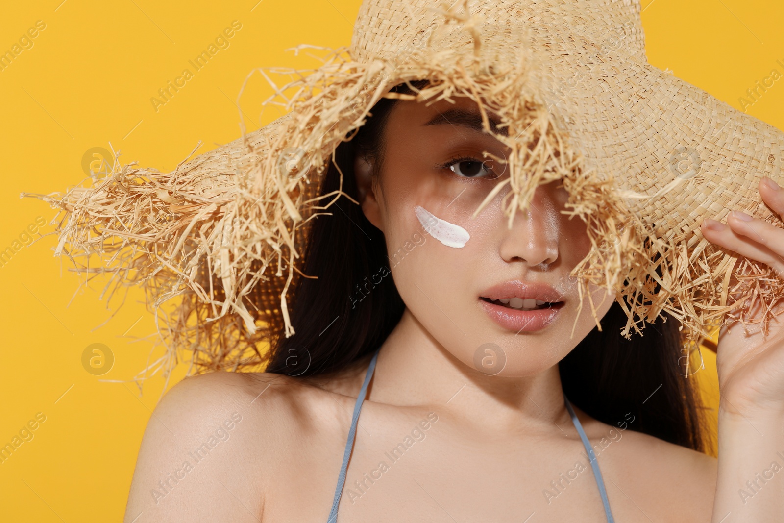 Photo of Beautiful young woman in straw hat with sun protection cream on her face against orange background