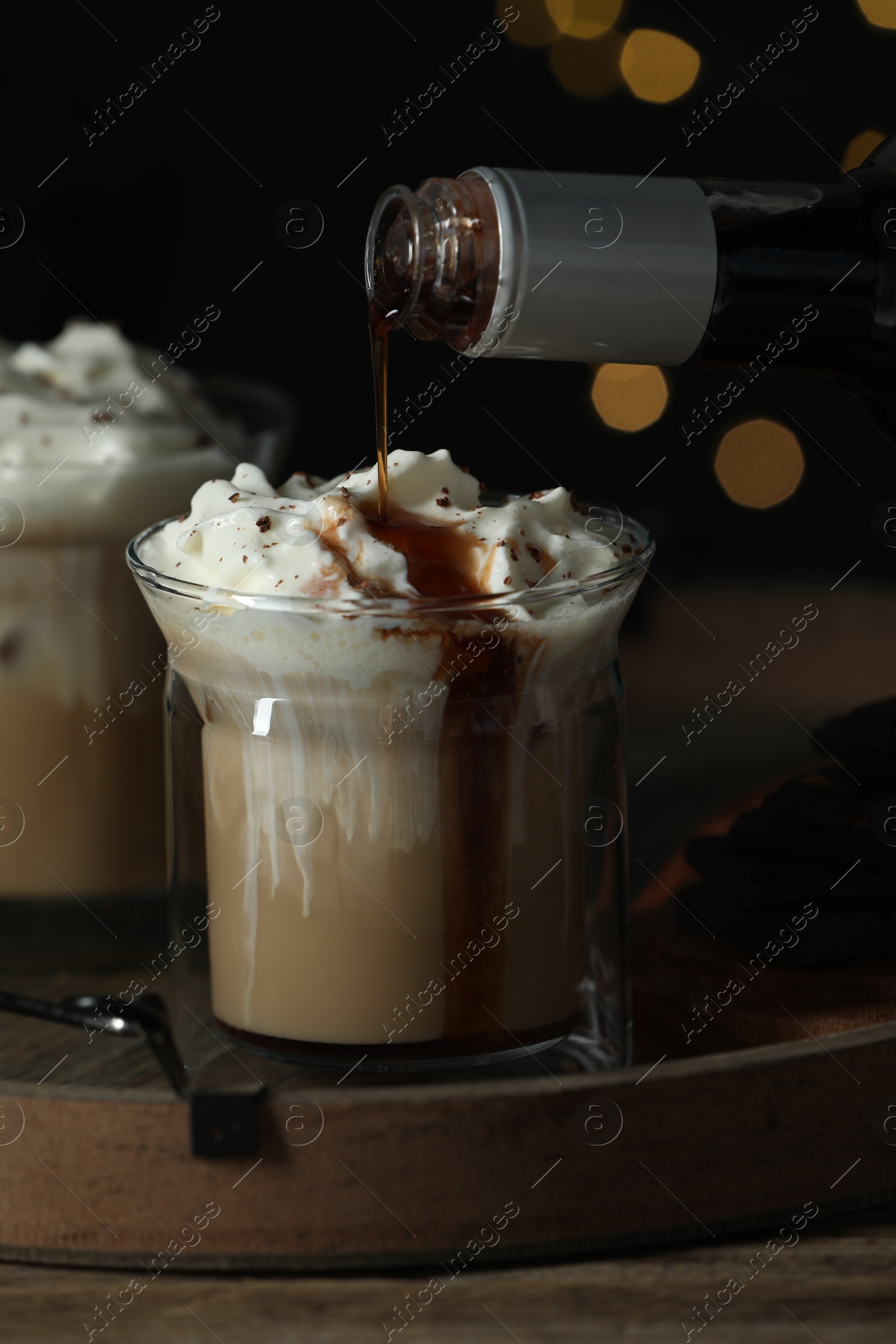 Photo of Pouring syrup into glass of tasty iced coffee against blurred lights on wooden table, closeup