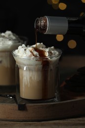 Photo of Pouring syrup into glass of tasty iced coffee against blurred lights on wooden table, closeup