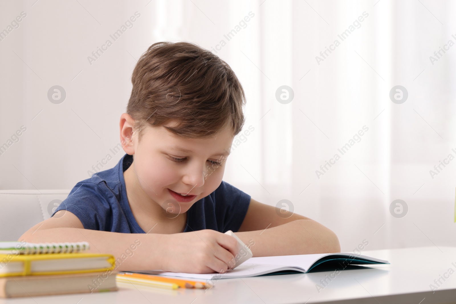 Photo of Little boy erasing mistake in his notebook at white desk indoors