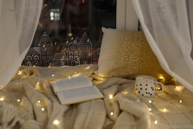 Open book and cup of hot drink on plaid near window with beautiful drawing at home. Christmas decor