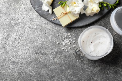 Body scrub in glass jar, soap bar and freesia flowers on grey table, flat lay. Space for text