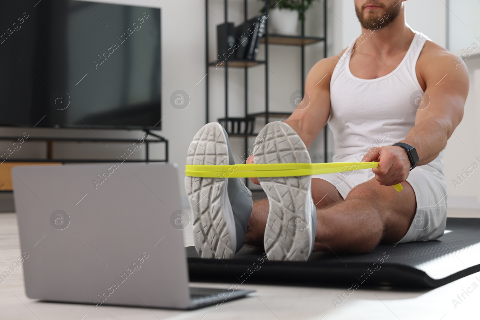 Photo of Muscular man doing exercise with elastic resistance band near laptop on mat at home, closeup