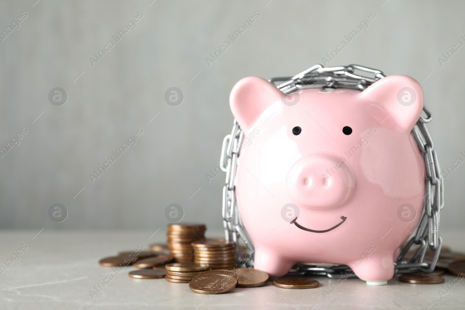 Photo of Piggy bank with steel chain and coins on grey marble table, space for text. Money safety concept