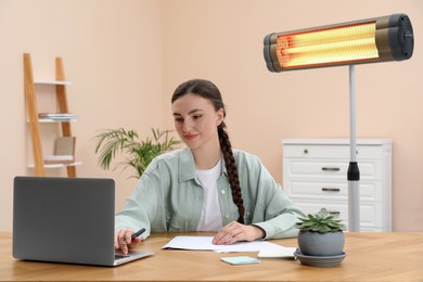 Photo of Young woman with laptop wooden table and modern electric infrared heater indoors