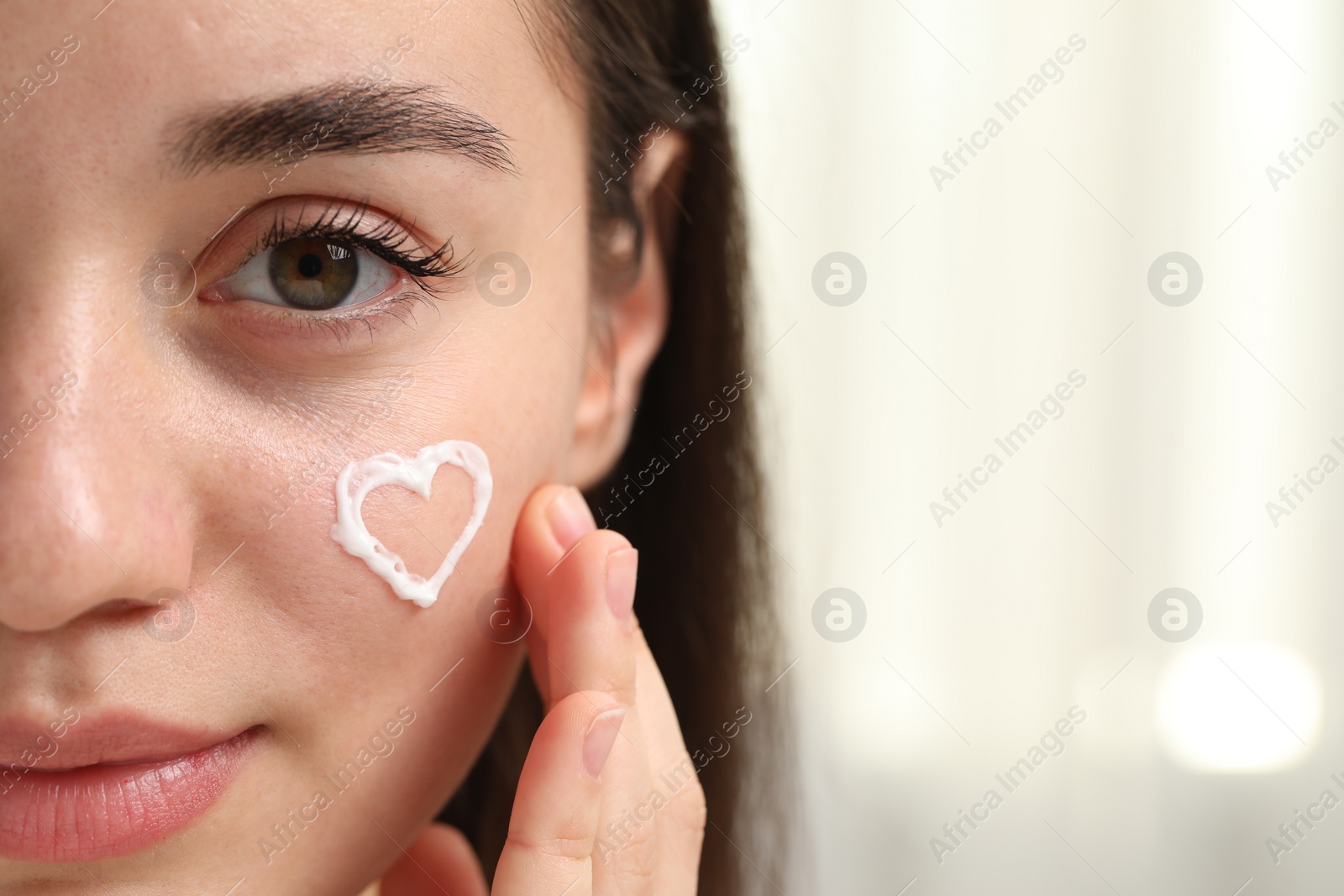 Photo of Young woman with dry skin and heart made of cream on her face indoors, closeup. Space for text