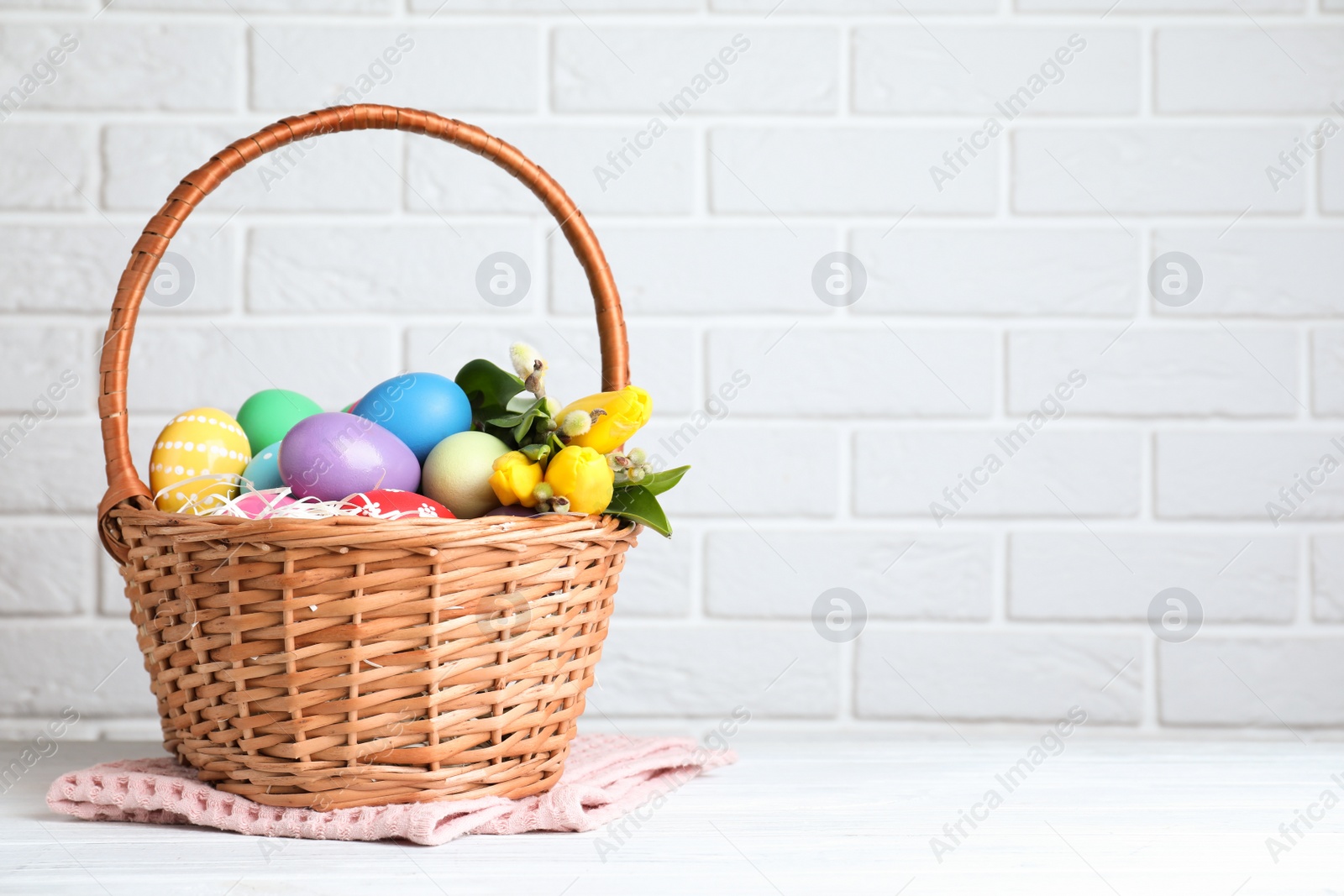 Photo of Wicker basket with bright painted Easter eggs and spring flowers on white wooden table near brick wall. Space for text