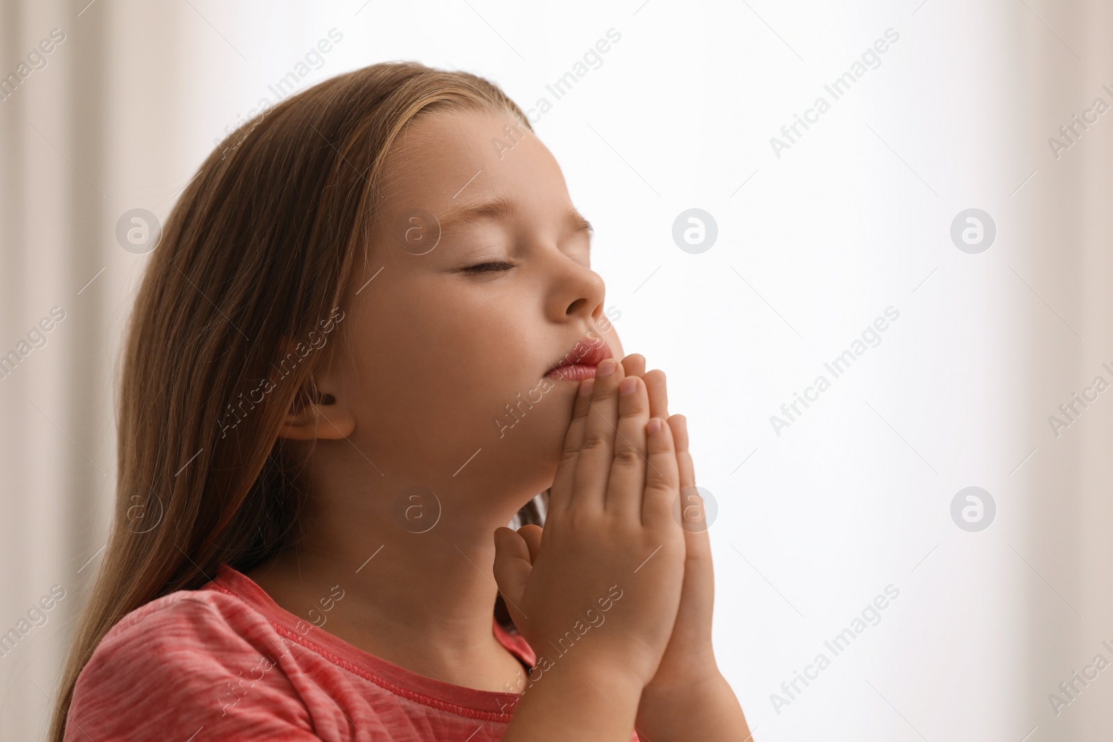 Photo of Cute little girl with hands clasped together praying on blurred background. Space for text