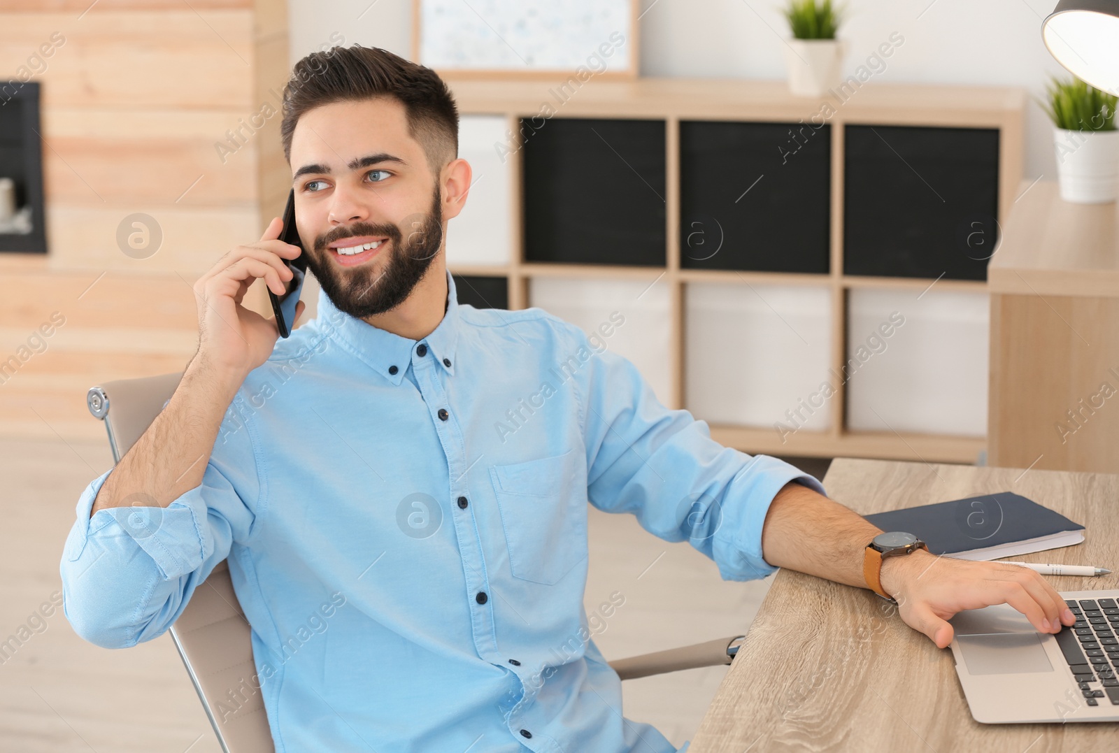 Photo of Handsome young man talking on phone while working at table with laptop in home office
