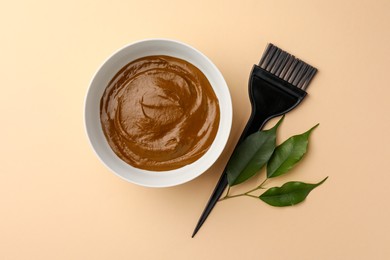 Photo of Bowl of henna cream, brush and green leaves on beige background, flat lay. Natural hair coloring