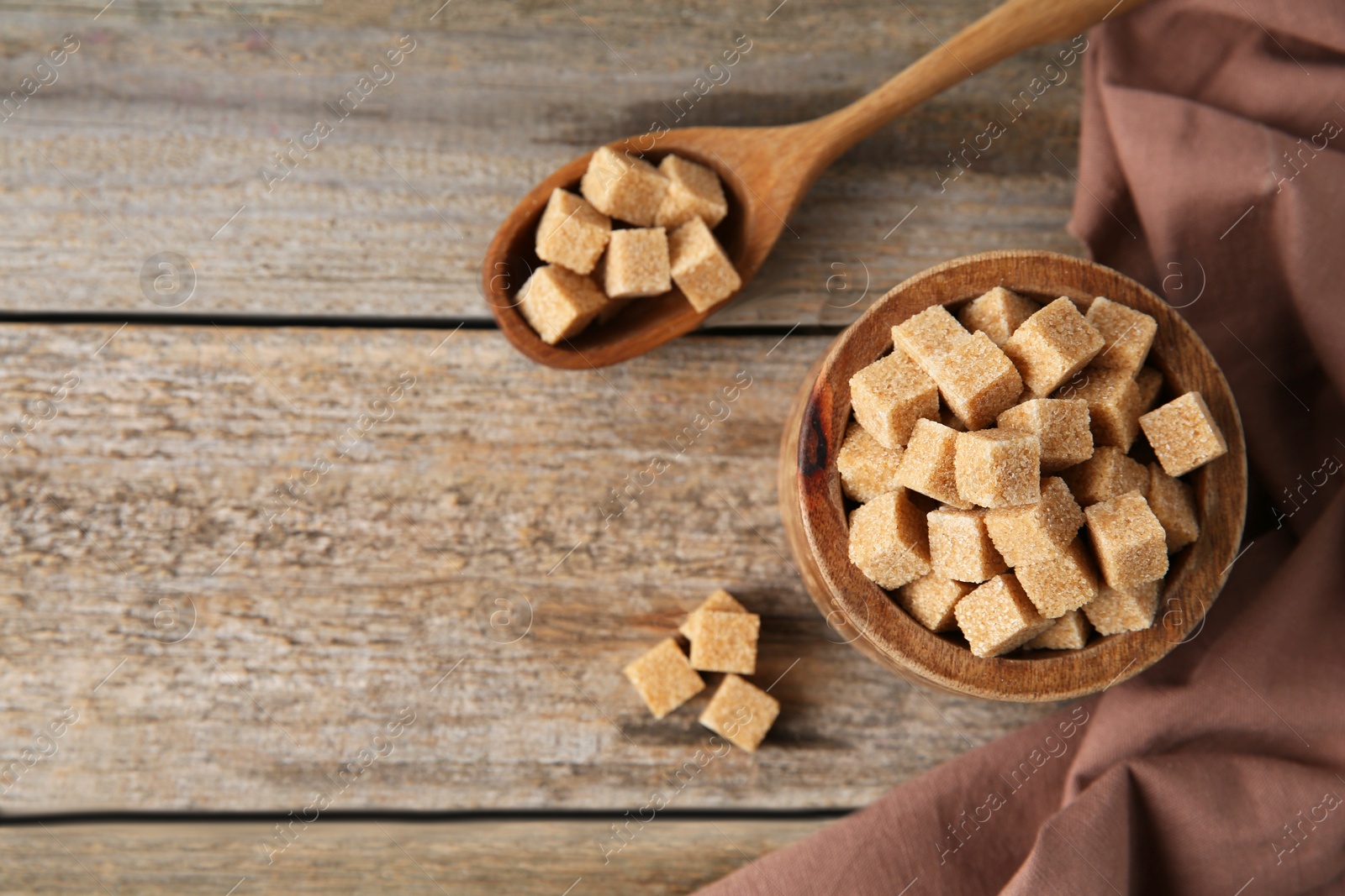 Photo of Bowl and spoon with brown sugar cubes on wooden table, flat lay. Space for text