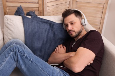 Upset man listening to music through headphones on sofa at home. Loneliness concept