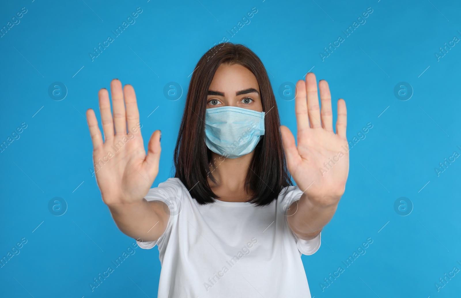 Photo of Young woman in protective mask showing stop gesture on light blue background. Prevent spreading of coronavirus