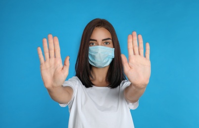 Photo of Young woman in protective mask showing stop gesture on light blue background. Prevent spreading of coronavirus