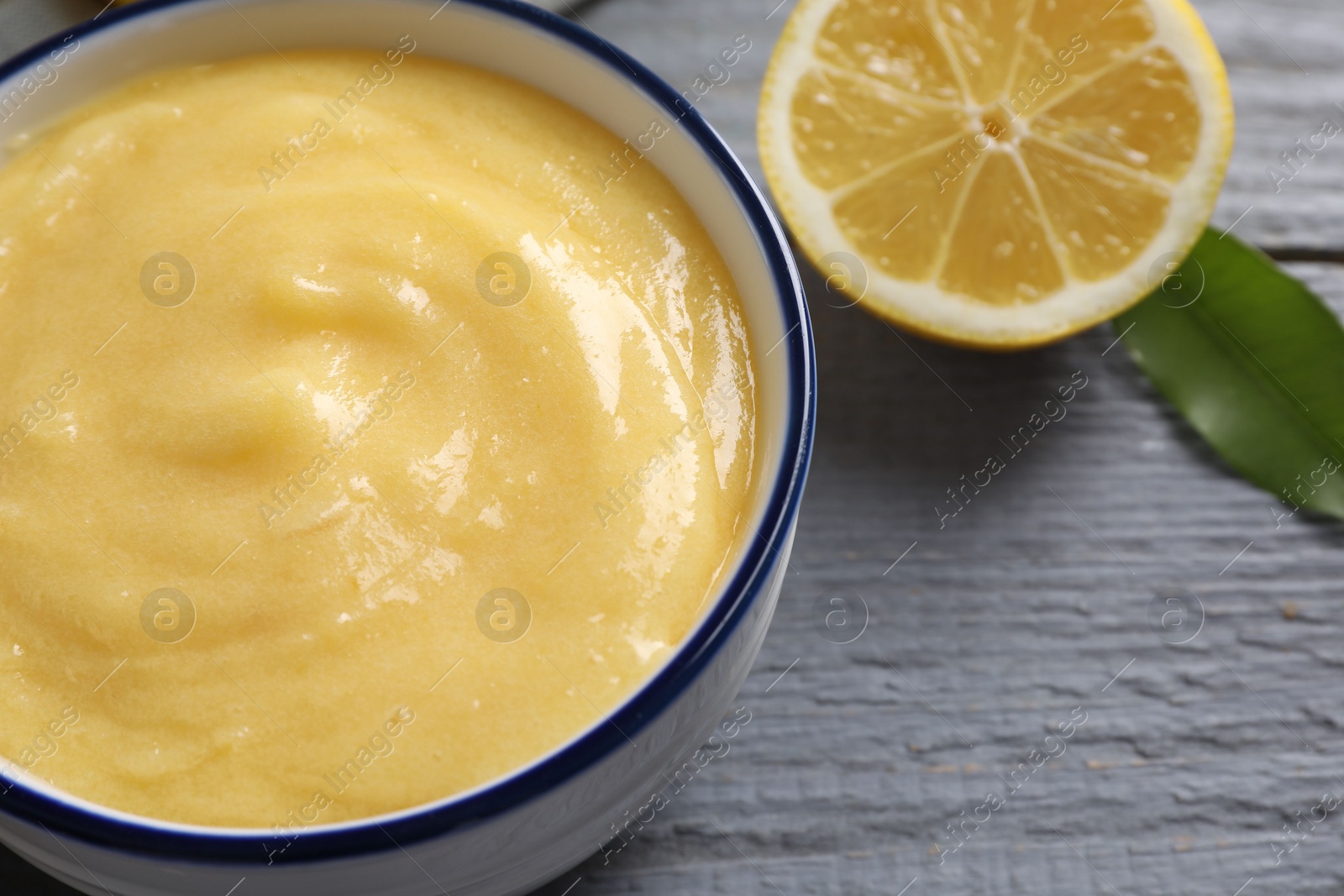 Photo of Delicious lemon curd in bowl, fresh citrus fruit and green leaf on grey wooden table, closeup