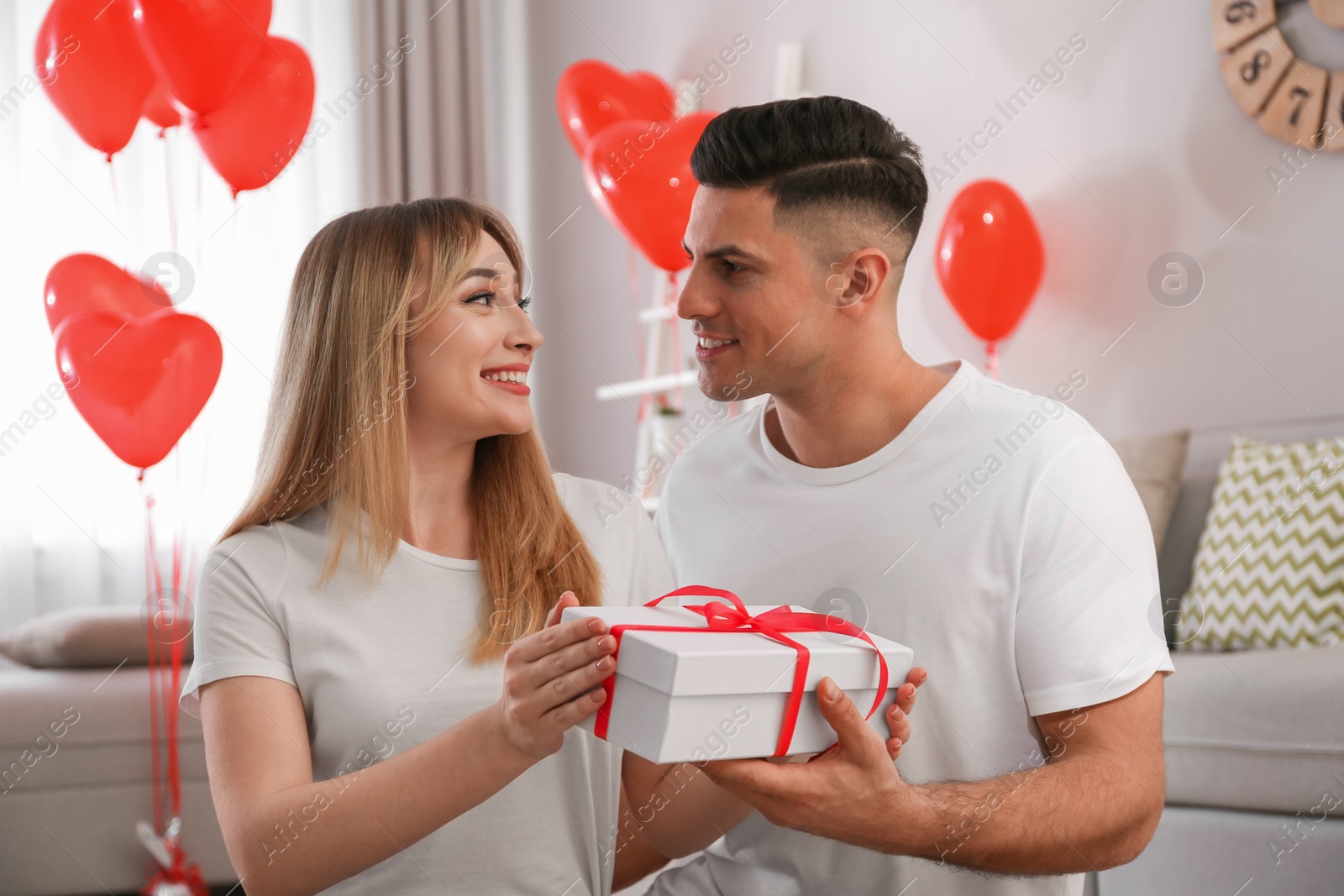 Photo of Man presenting gift to his girlfriend in room decorated with heart shaped balloons. Valentine's day celebration