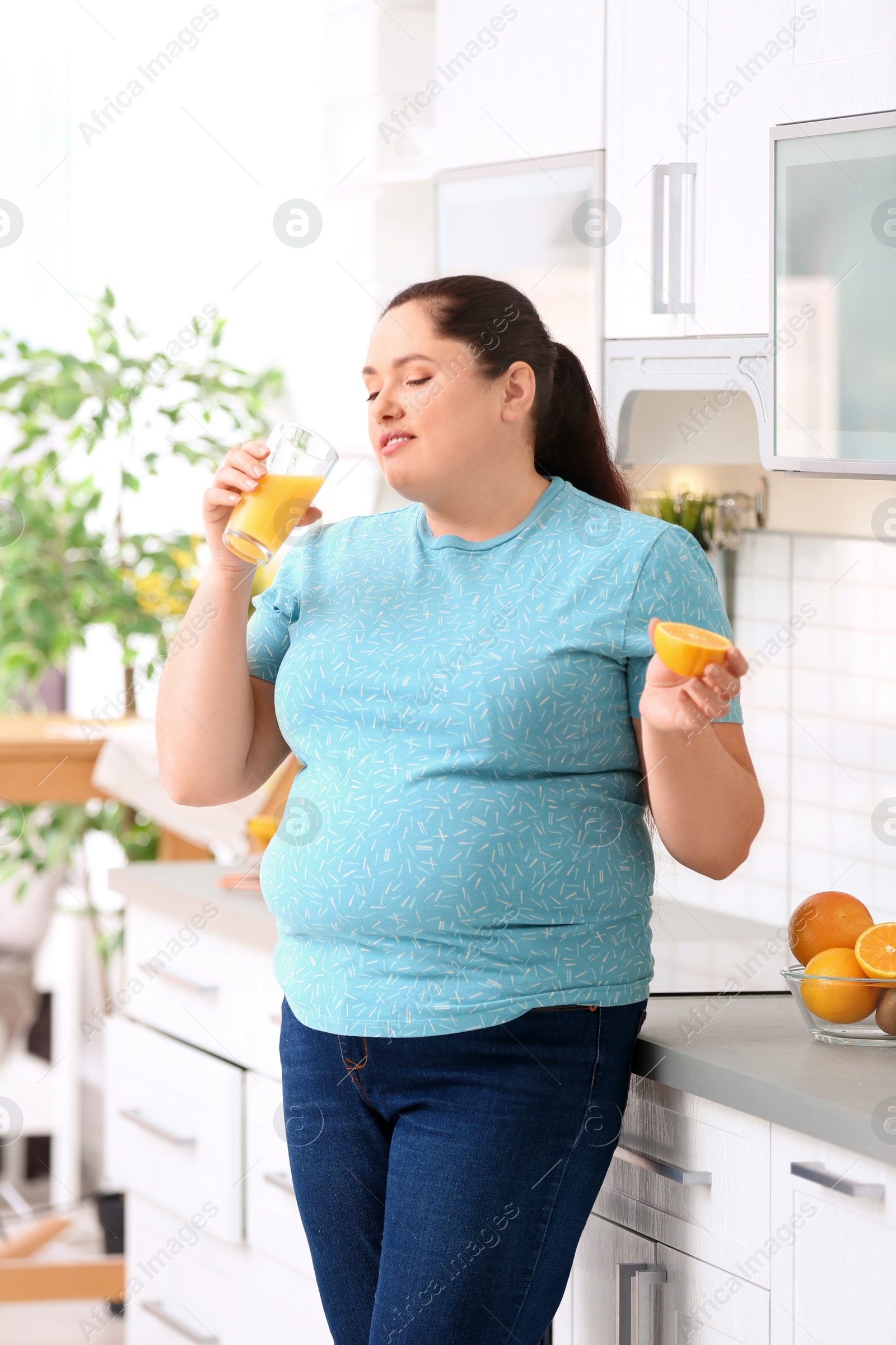 Photo of Overweight woman drinking fresh juice and orange in kitchen. Healthy diet