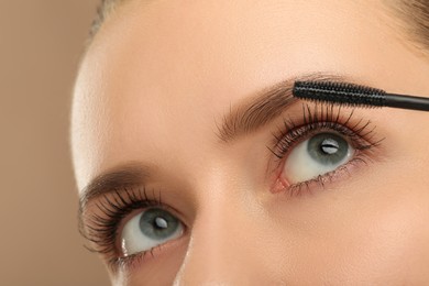 Photo of Woman applying mascara onto eyelashes against light brown background, closeup