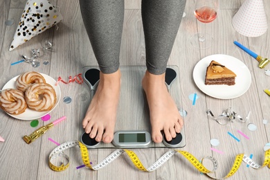 Photo of Woman using scale surrounded by food and alcohol after party on floor. Overweight problem