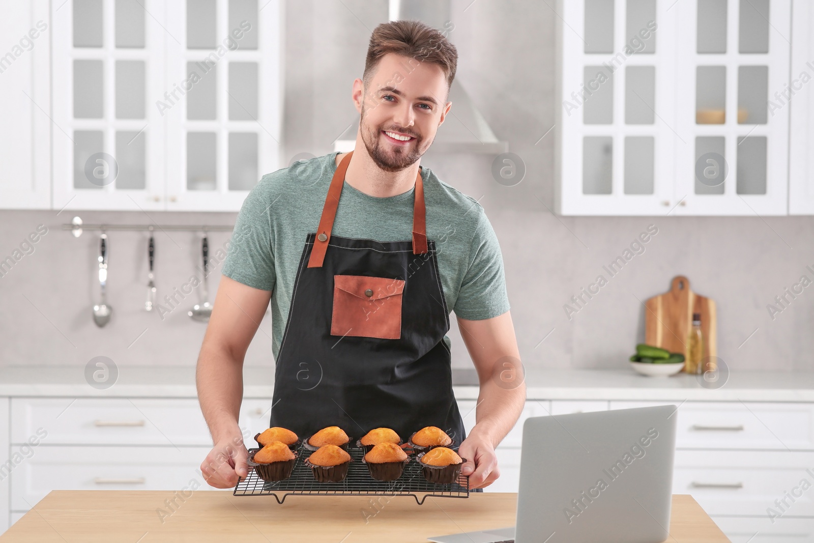 Photo of Happy man holding muffins near laptop at table in kitchen. Time for hobby