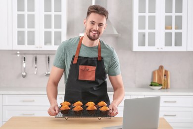 Photo of Happy man holding muffins near laptop at table in kitchen. Time for hobby
