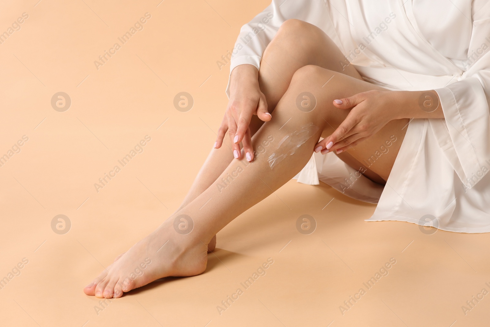 Photo of Woman applying body cream onto her smooth legs on beige background, closeup