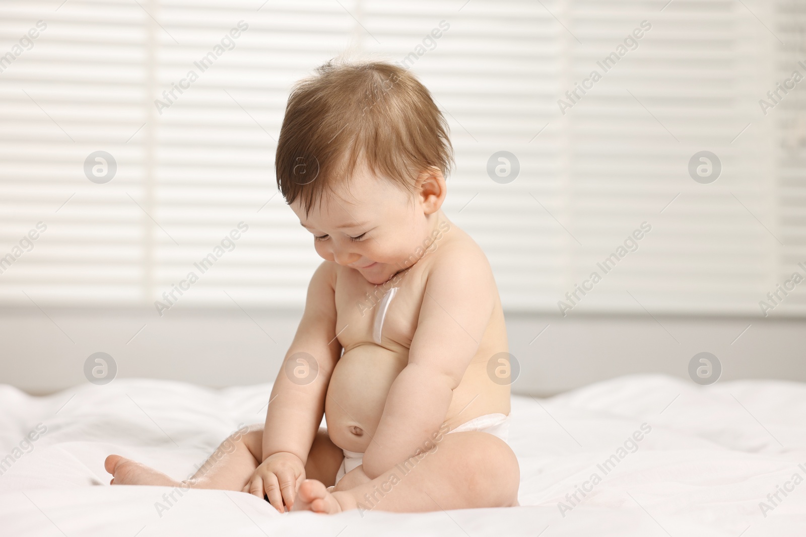 Photo of Cute little baby with moisturizing cream onto body sitting on bed at home