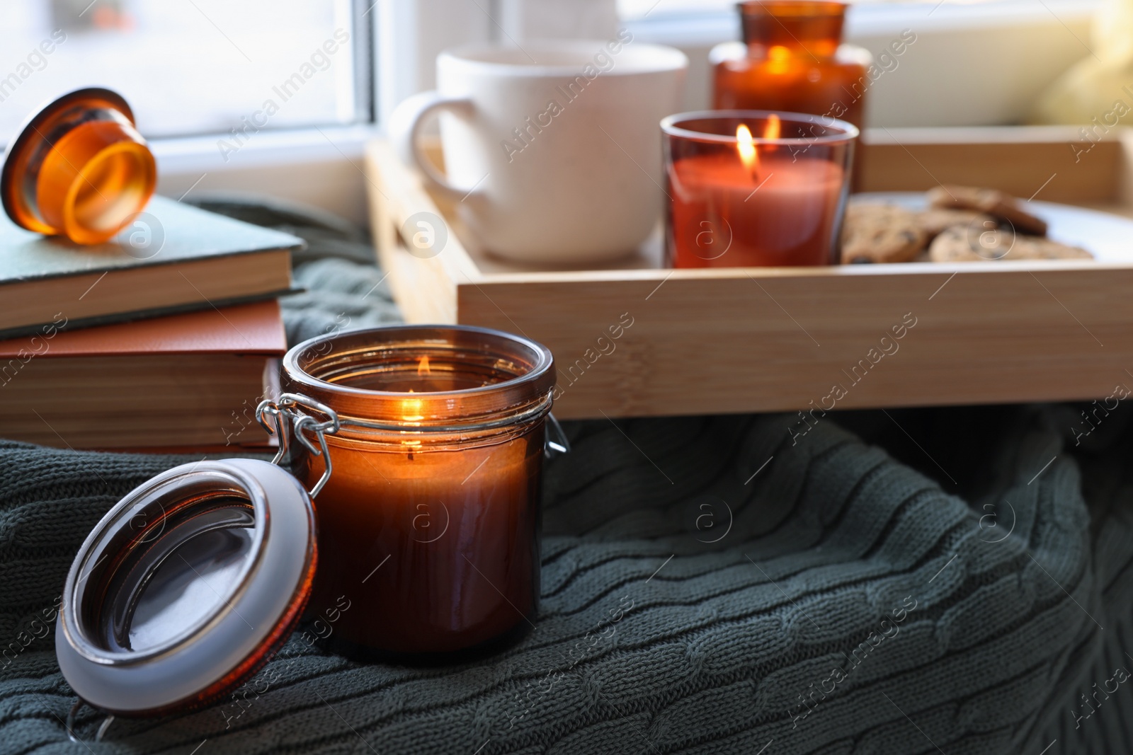 Photo of Burning candle in jar on knitted plaid near window indoors
