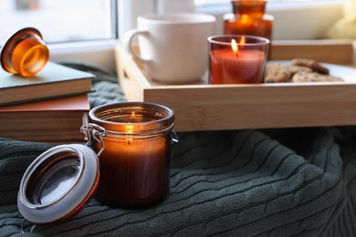 Burning candle in jar on knitted plaid near window indoors