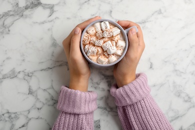 Photo of Woman in sweater holding cup of tasty chocolate with milk and marshmallows on marble table, top view